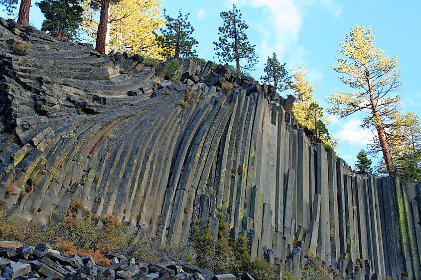 Curved basalt columns form a striking cliff at Devil's Postpile in California. Pine trees crown the formation, under a partly cloudy blue sky.