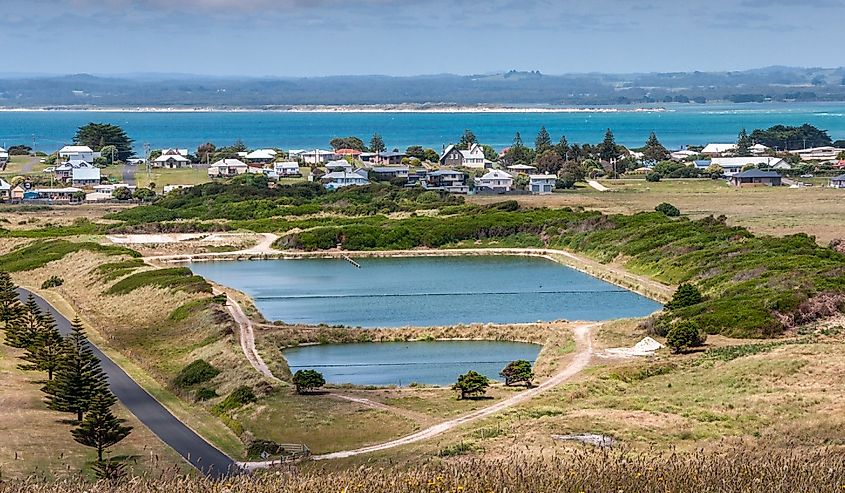 Fresh water reservoir slightly above the white houses of the town with ocean in back. Mountains under cloudscape on horizon.