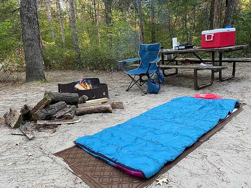 A campsite with a sleeping bag laid out on the ground, a picnic table, and a burning fire pit.