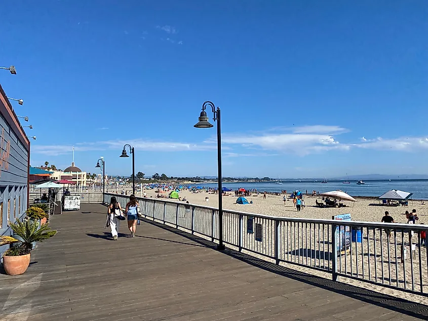 Two girls walk along a beach boardwalk, with lots of beachcombers to the right and a large carnival in the distance