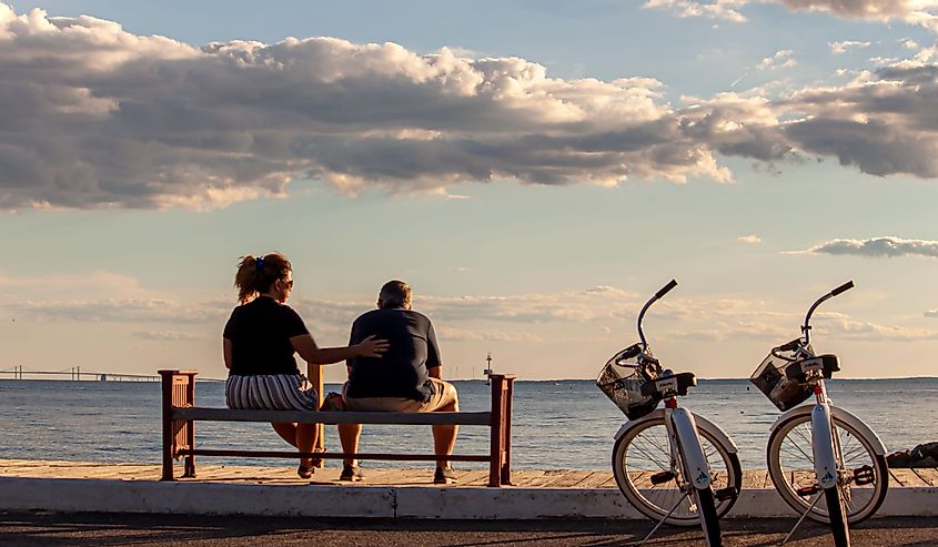 A middle aged caucasian couple is sitting on a bench by the beach. They have their identical bikes parked next to them. Woman gives the man a pat on the back, Rock Hall, Maryland