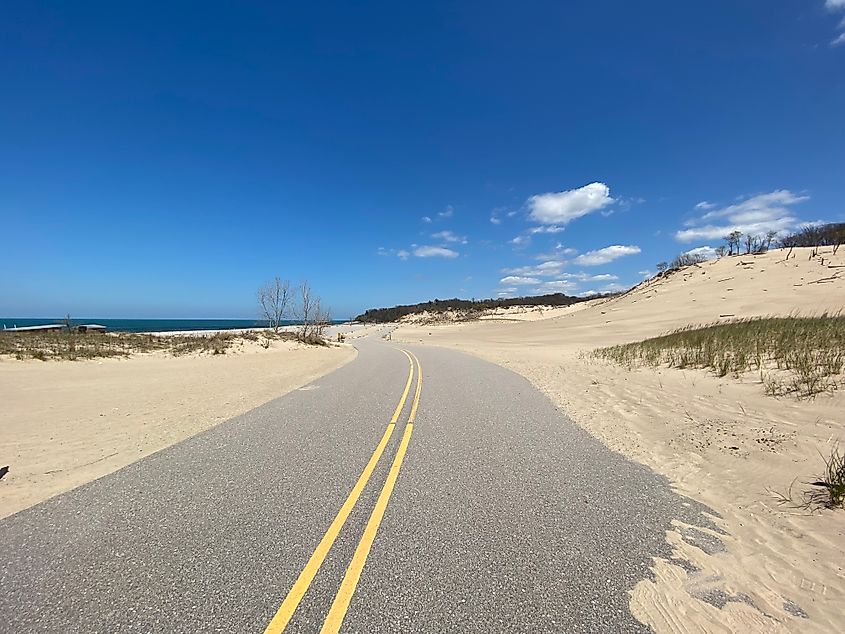 A two-lane, sand-swept road cuts between a large sand dune and Lake Michigan beach.