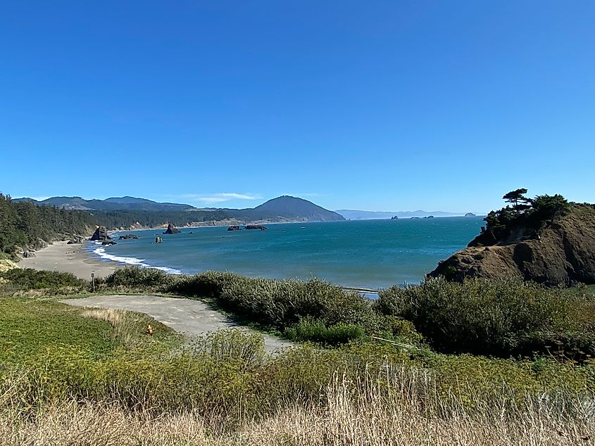 A sweeping view of windswept grass, rock monoliths, a long flat beach, and the Pacific Ocean on a bluebird day. 
