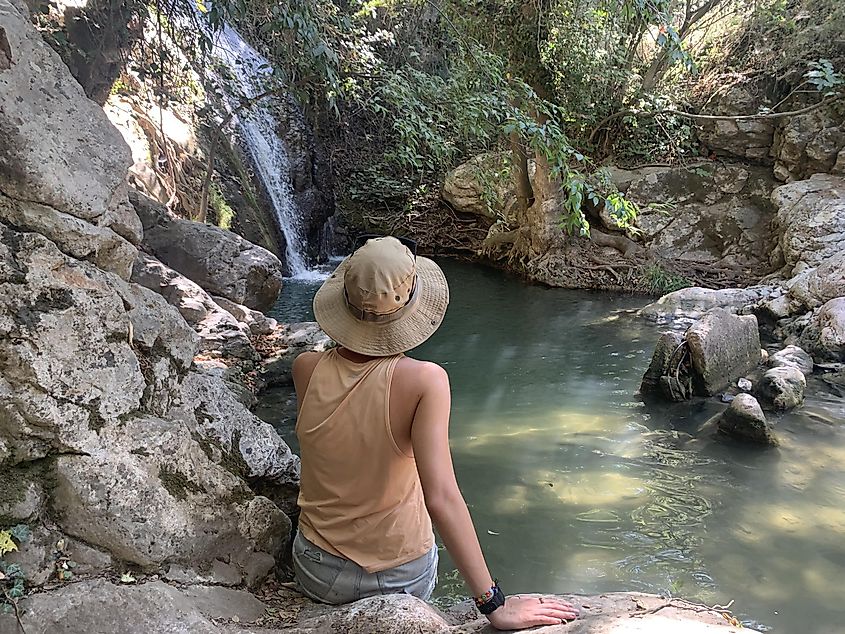 A woman sits by a small natural pool at the base of a waterfall in the forest.