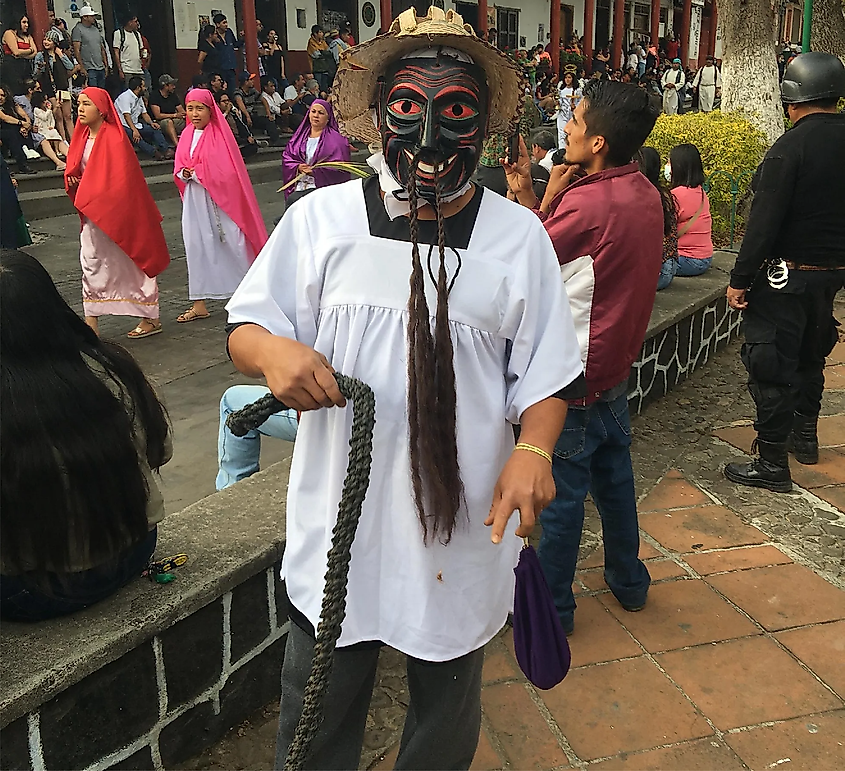 A man in a devilish mask holds a whip while a parade marches on in the background.