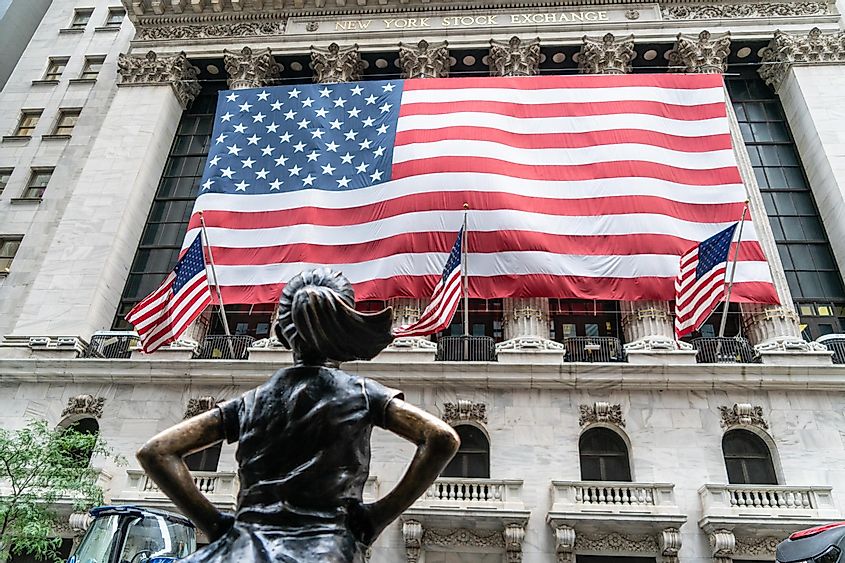 Fearless Girl Standing Bronze Strong Looking Up At The Iconic Structure Of The NY Stock Exchange In Lower Manhattan With American Flag.