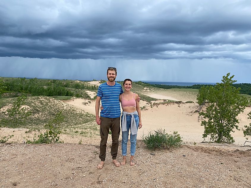 A couple poses atop a large sand dune complex as rain clouds approach from behind.