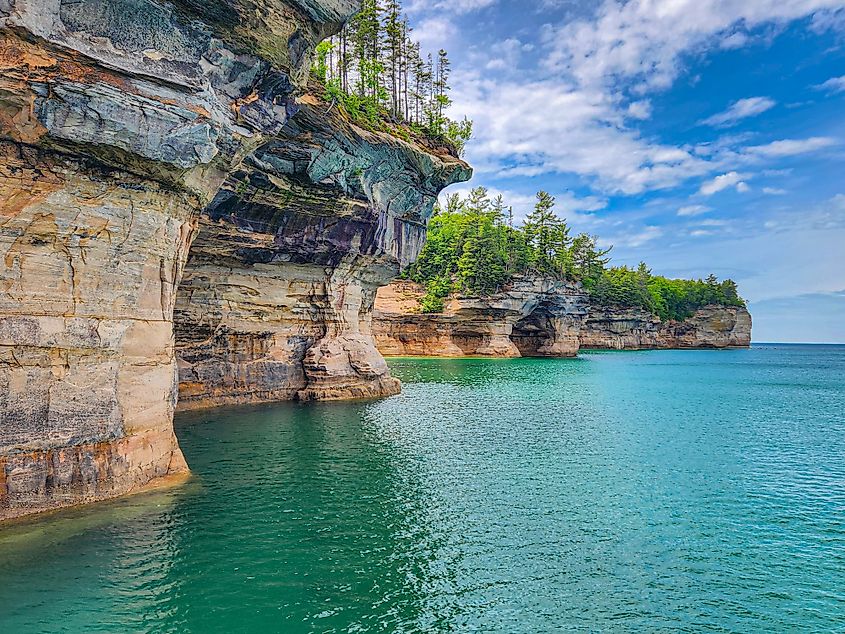 Layered sandstone cliffs rise above clear turquoise water at Pictured Rocks in Michigan, with pine trees growing along the cliff tops under a partly cloudy blue sky.