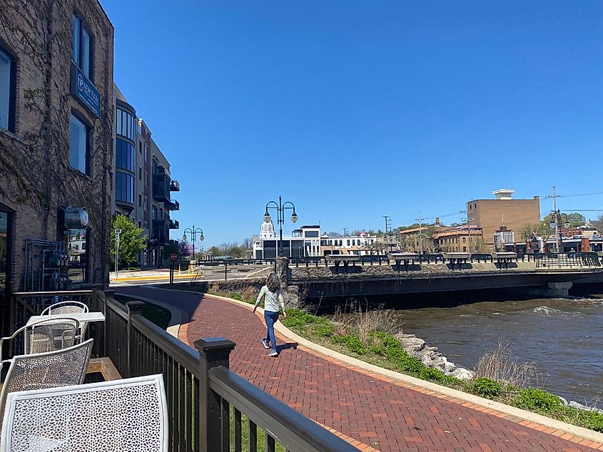 A woman walks a cafe patio via a brick-lined riverside path.