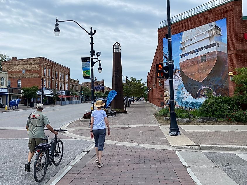 A couple walks across a crosswalk, into a red brick downtown with a large mural of a lake freighter.