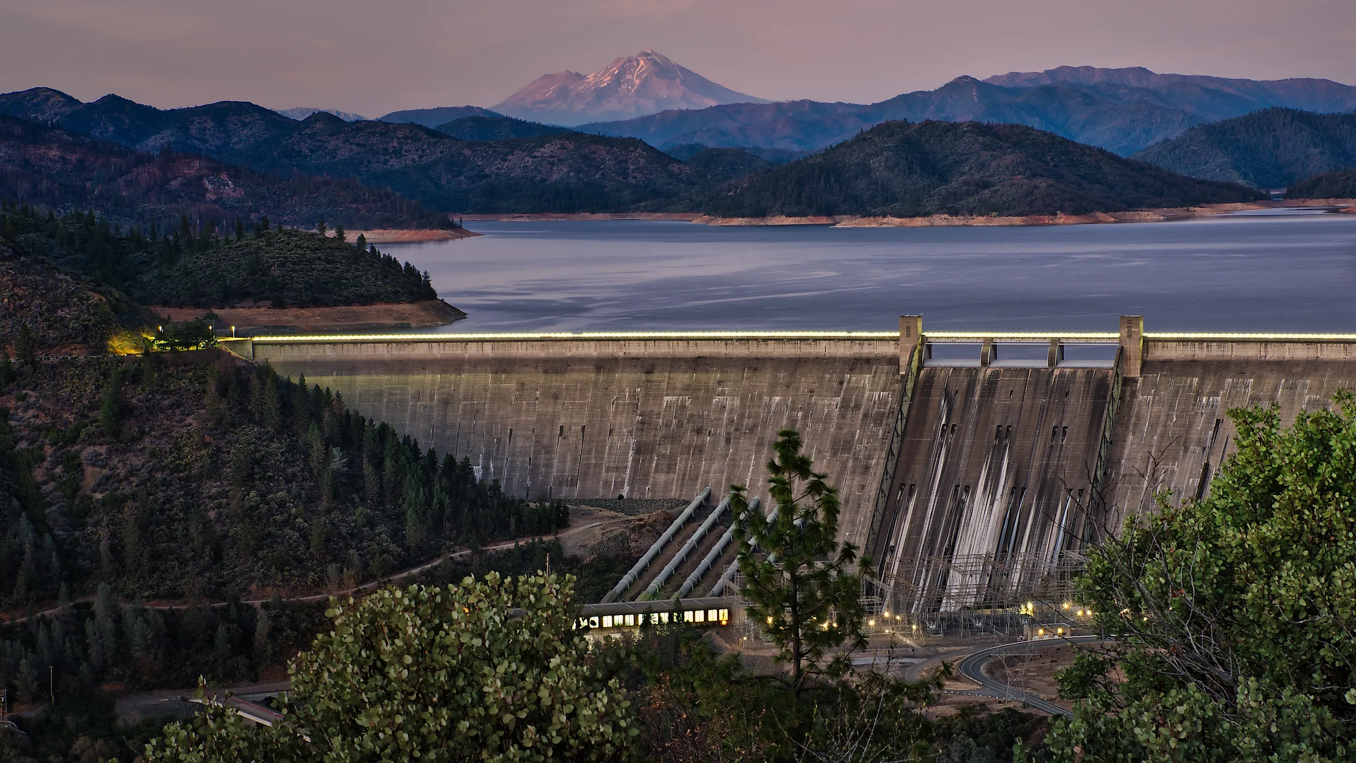 A picture of Shasta Dam with a lake and rocky mountains on the background during sunset. 
