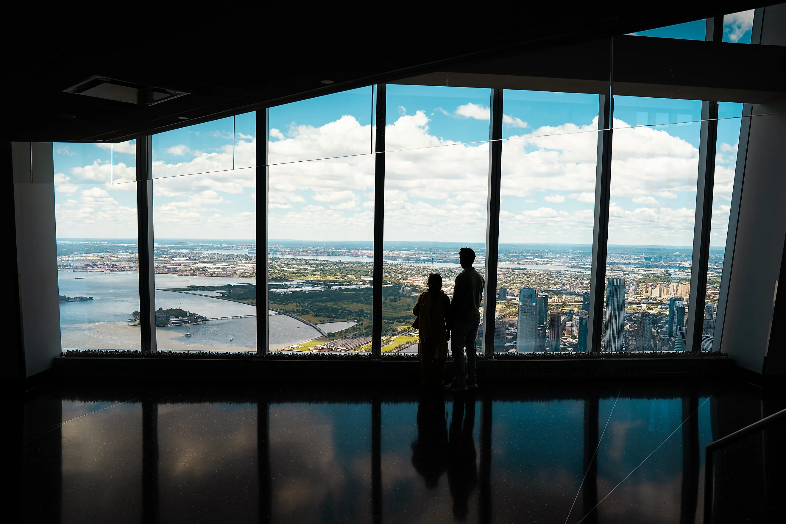Couple standing in front of the large glass windows at One World Observatory, overlooking the New York City
