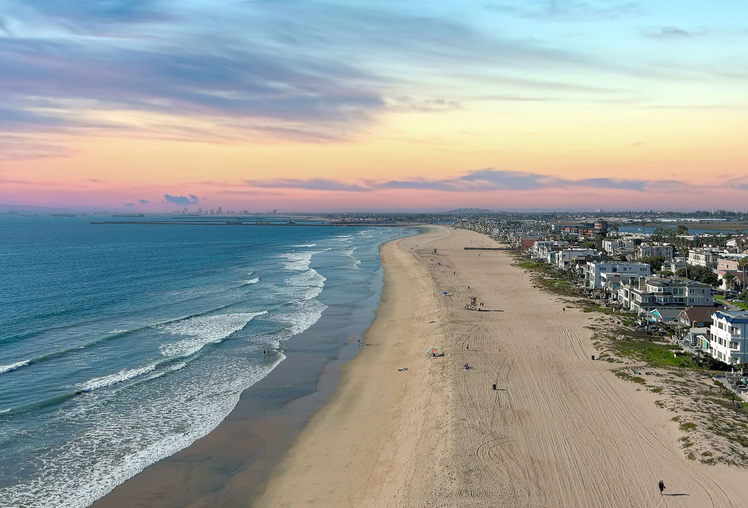 Aerial shot of the coastline with blue ocean water and homes along the sand on the beach, and blue sky with clouds in Huntington Beach California.