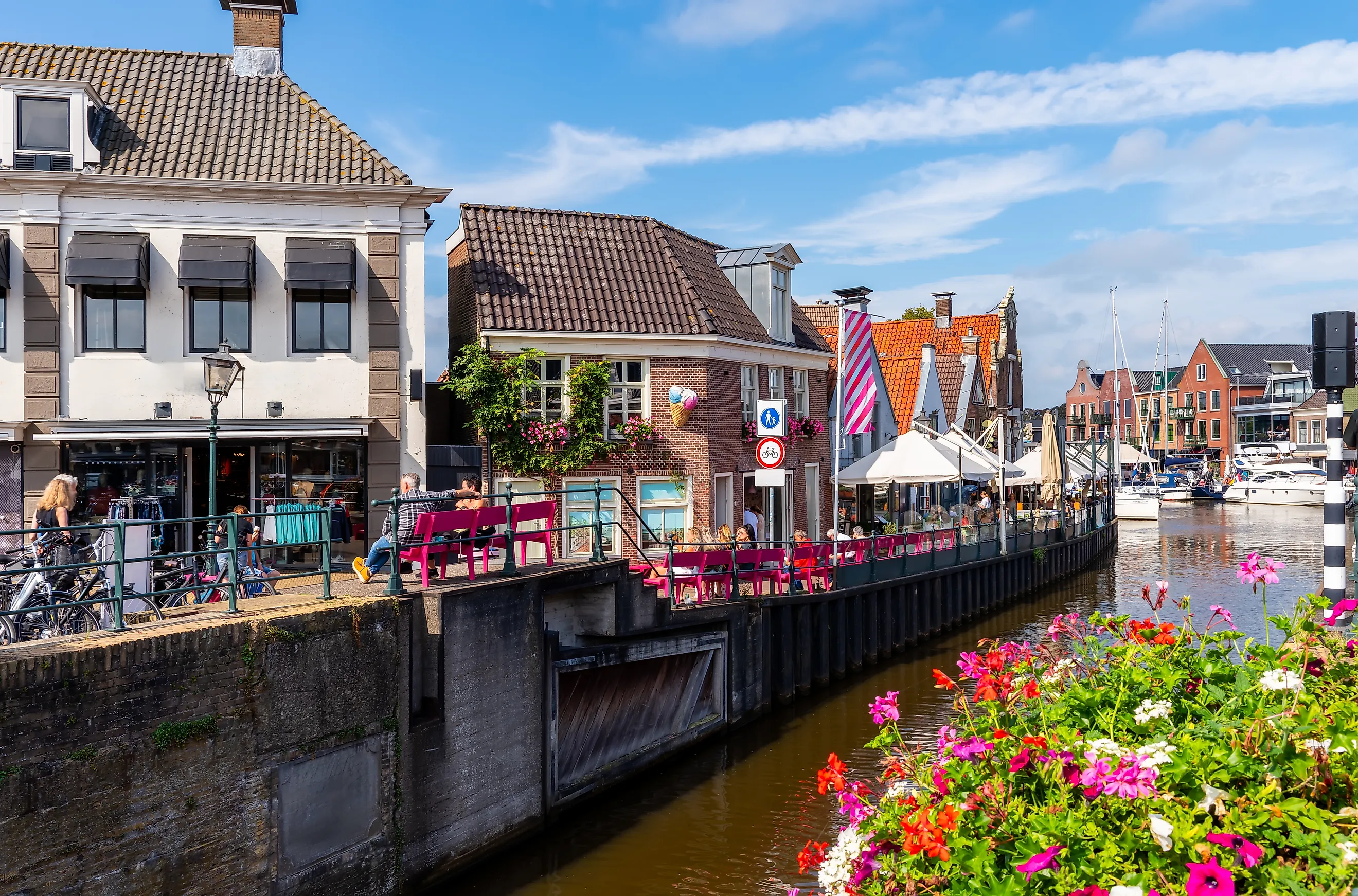 A vibrant canal-side scene in Lemmer, Netherlands, featuring historic Dutch buildings, a cozy café with pink benches, and colorful flowers.