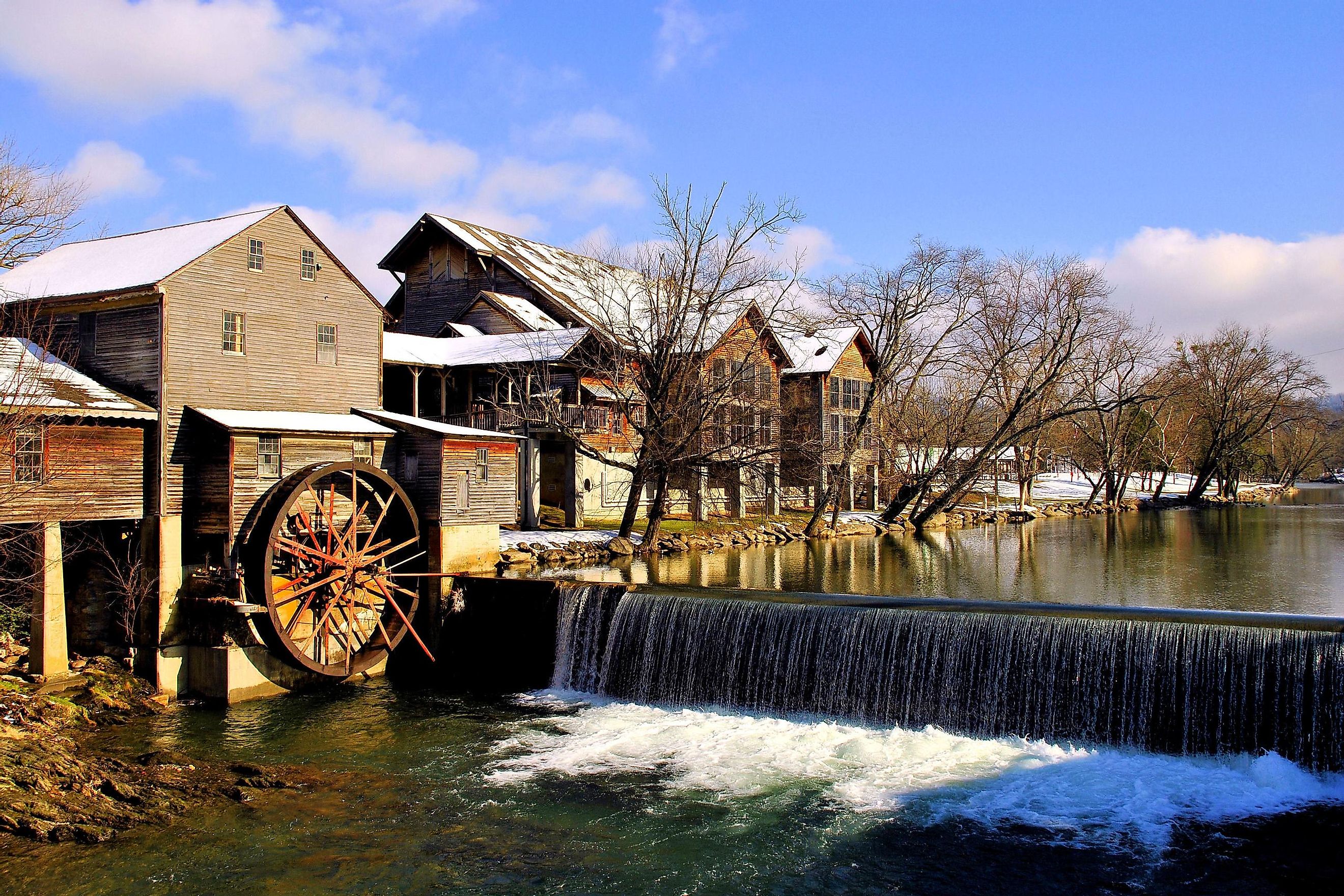 The Old Mill in Pigeon Forge, Tennessee, surrounded by snow, with a picturesque waterwheel and the Smoky Mountains in the background.