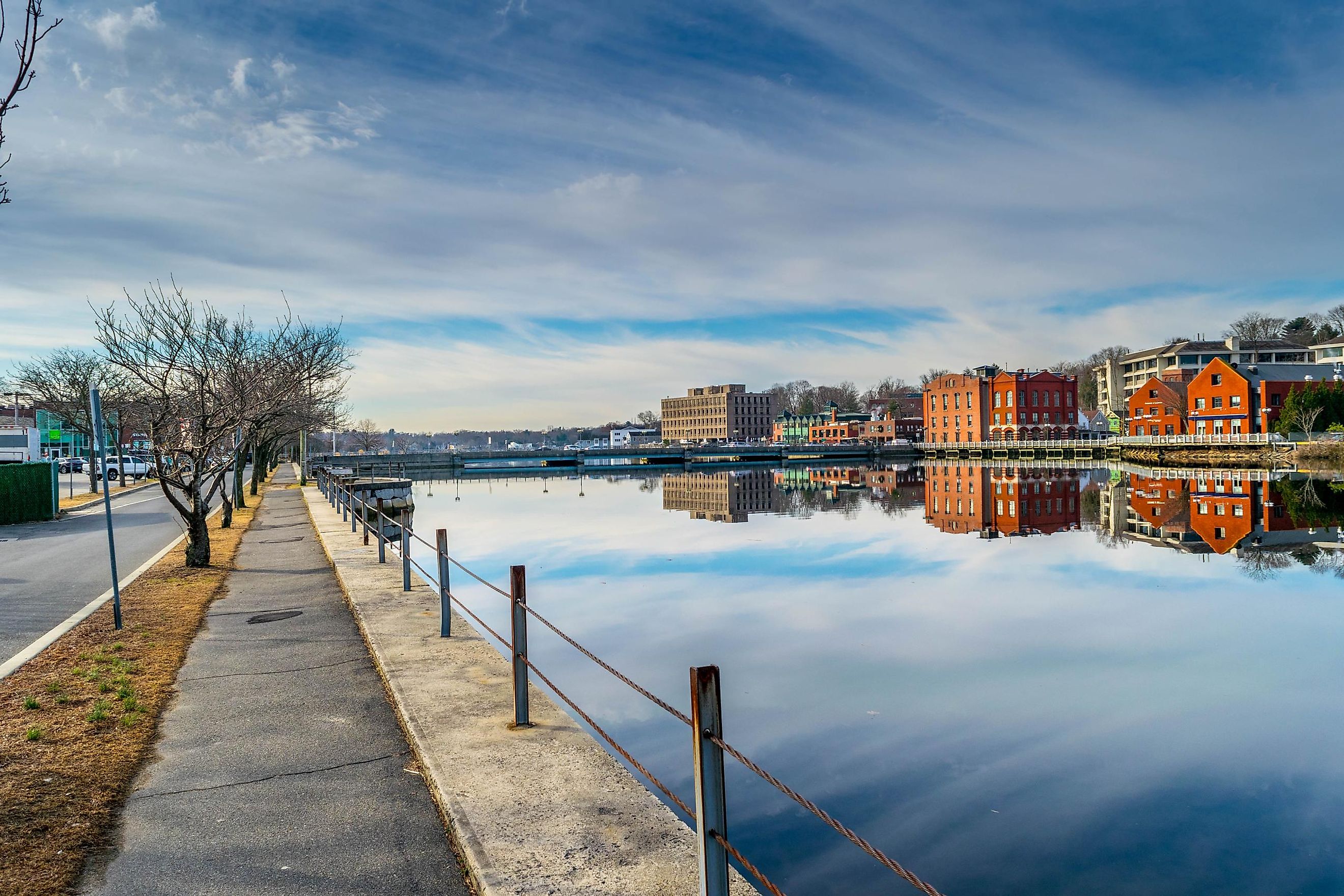 A serene view of Westport, Connecticut's downtown area, with calm waters reflecting colorful buildings, a clear blue sky overhead, and a paved walkway lined with bare trees.