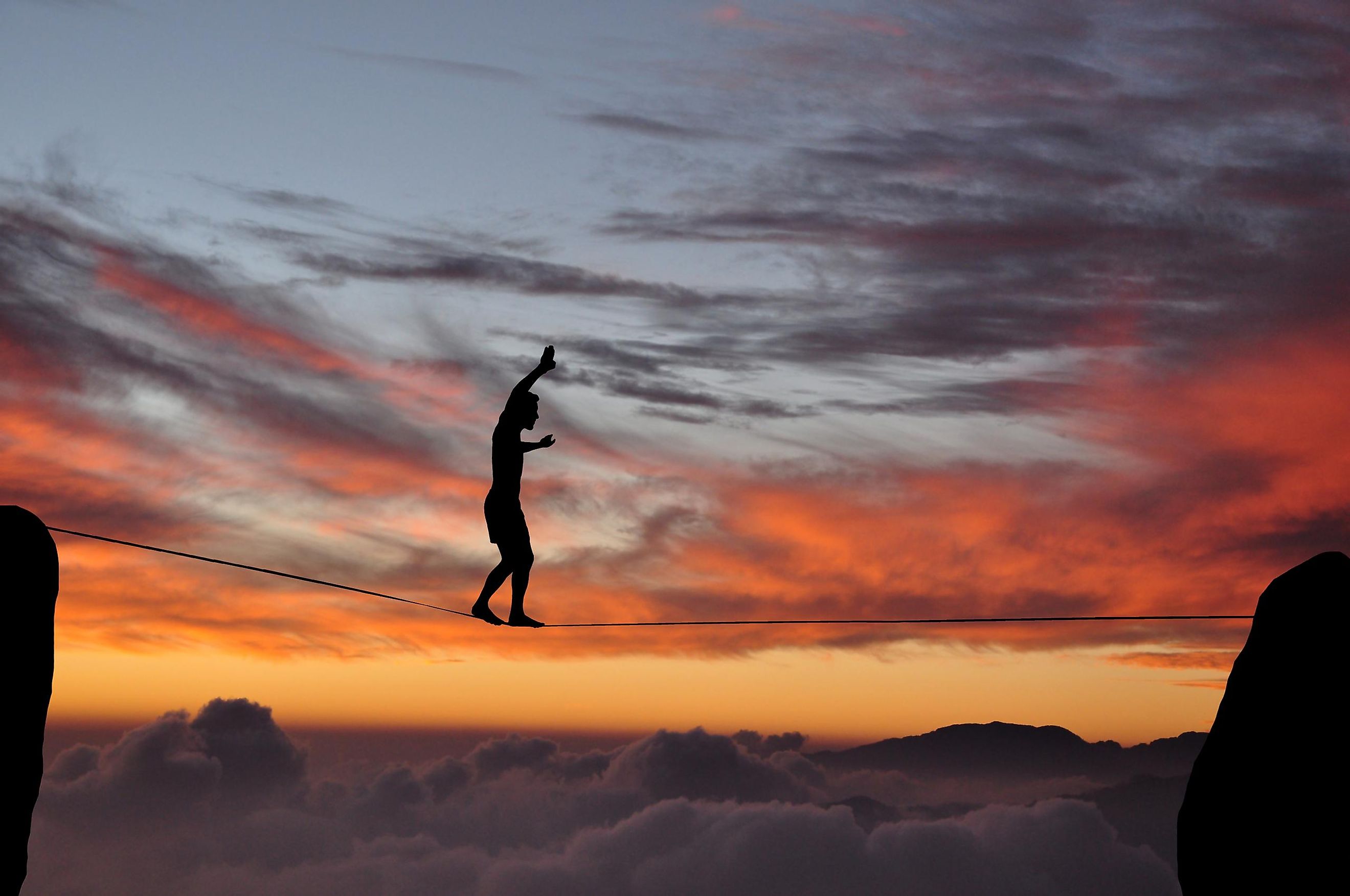 Silhouette of young man balancing on slackline high above clouds and mountains during sunset