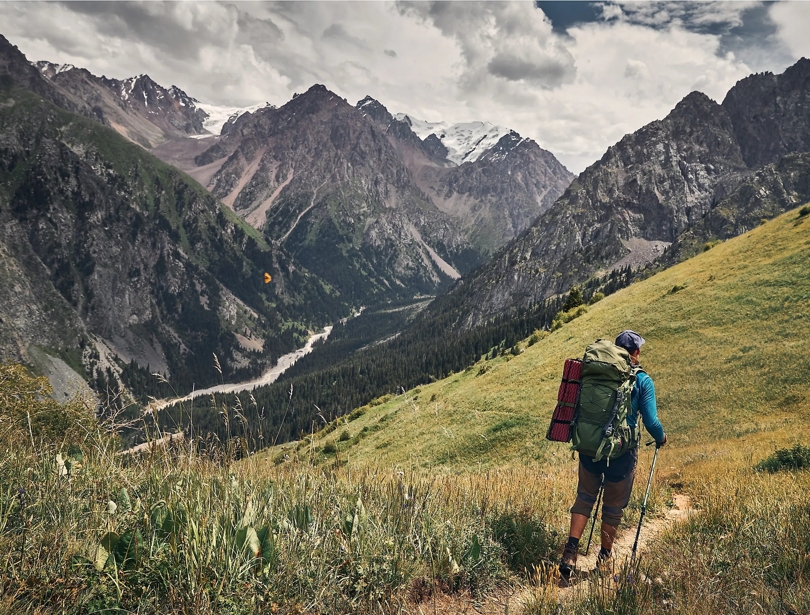 Hiker walking in the green mountain valley with snowy peaks and cloudy sky.