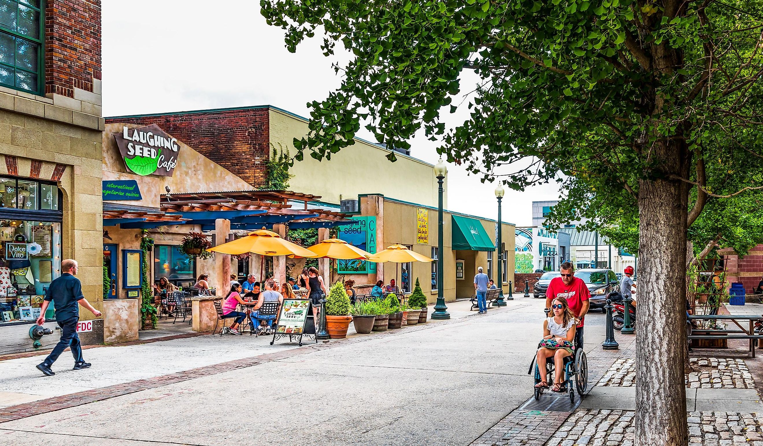 Wall Street in downtown is busy on a summer Sunday, with small crowds at cafe tables, while a man pushes a young woman in a wheel chair.