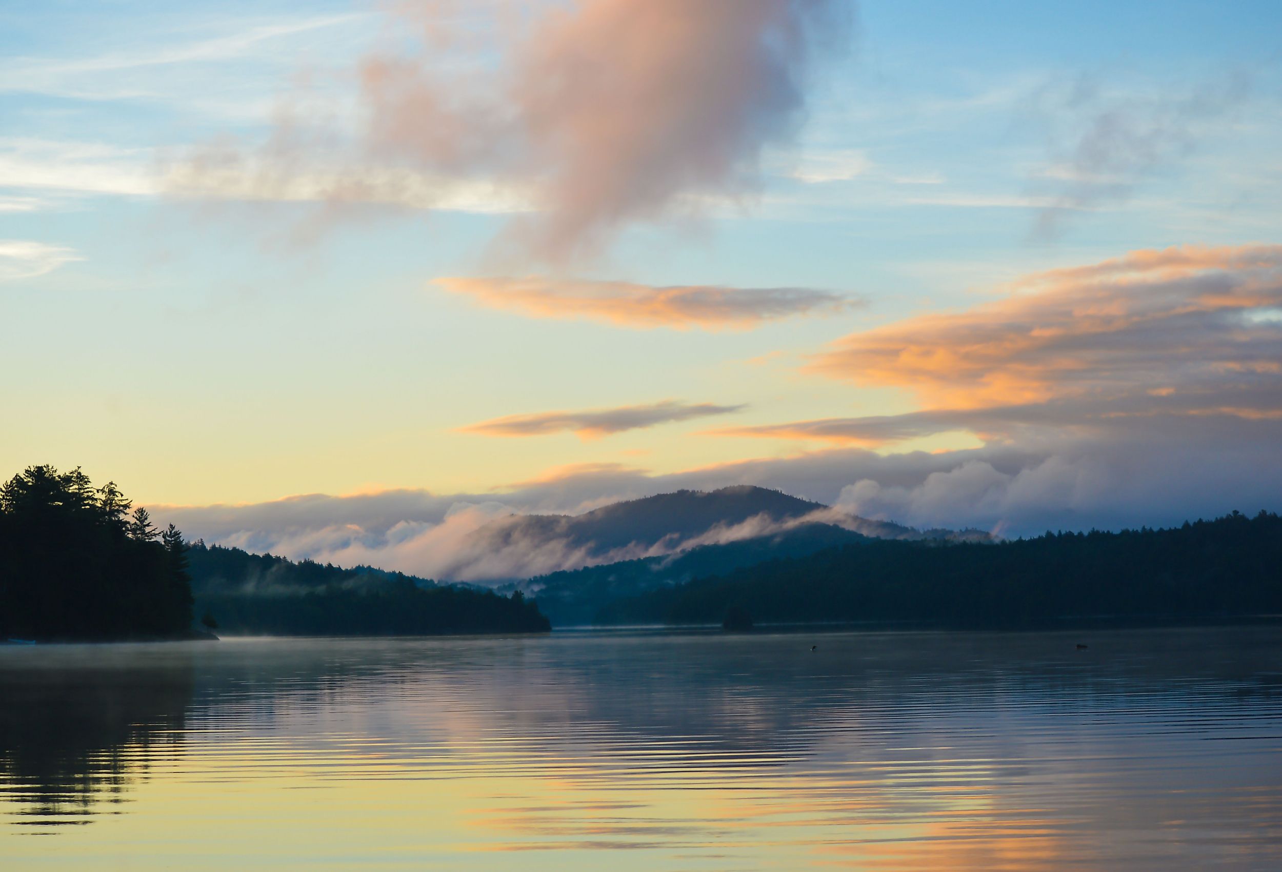 Sun rising over Saranac Lake with the Adirondack Mountains and rolling fog reflecting in the lake; two loons float on the water.