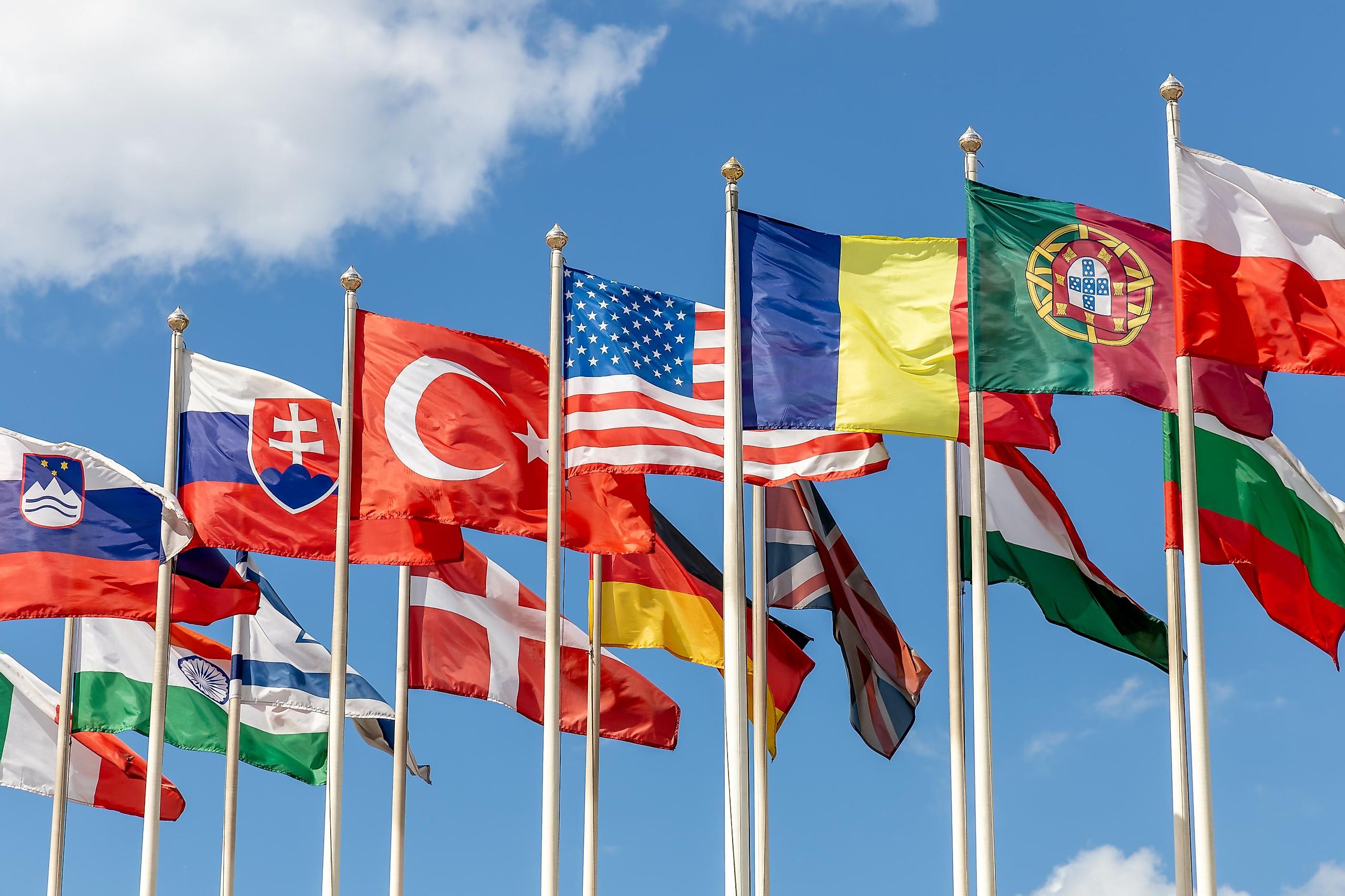 A group of flags from countries from around the world on flagpoles, fluttering in the wind against the backdrop of a blue sky.