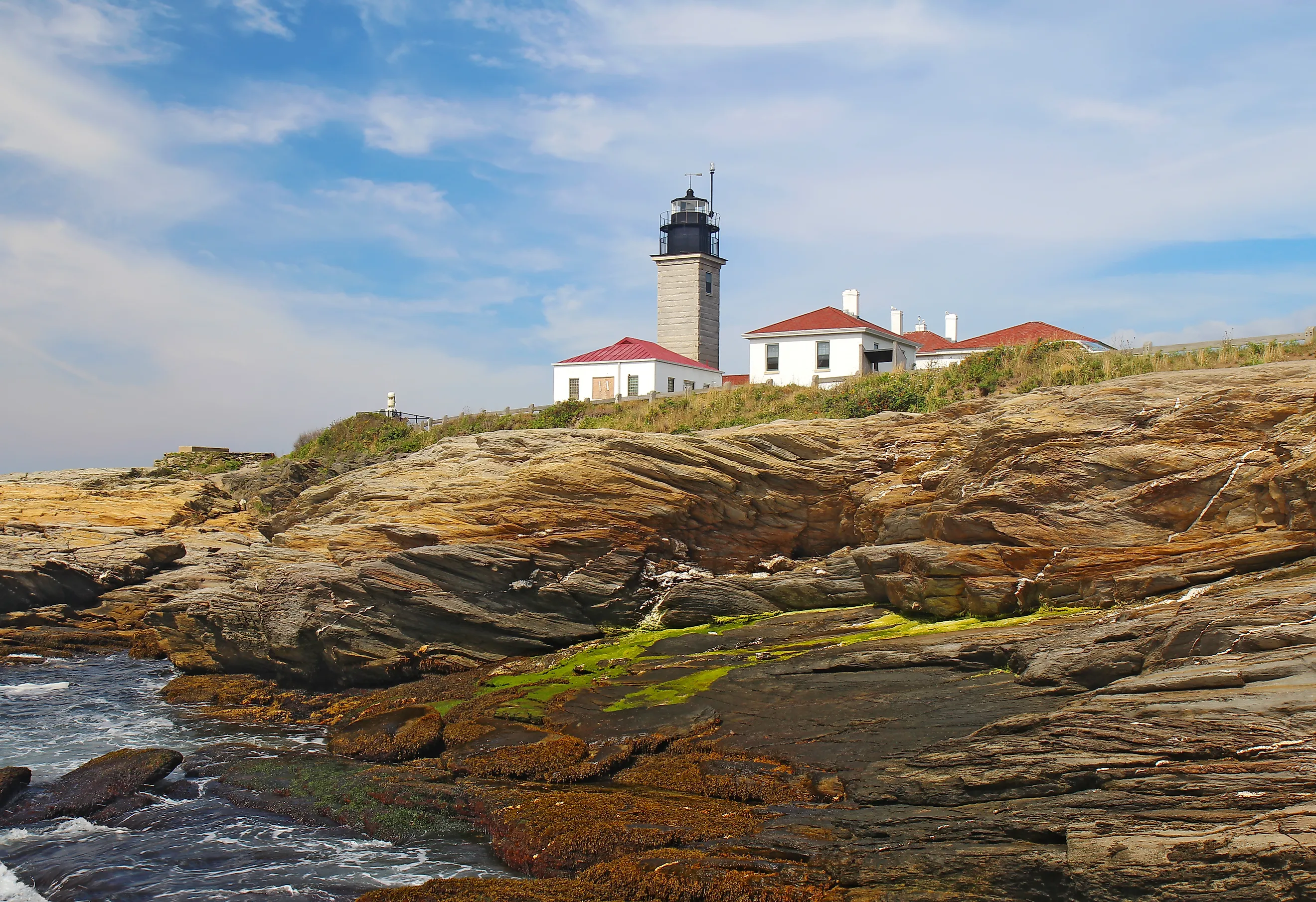 Editorial Photo Credit:Stephen B. Goodwin via Shutterstock. The Beavertail Light lighthouse near Jamestown on Conanicut Island, Rhode Island, viewed from the rocky coast with a bright blue sky and white clouds