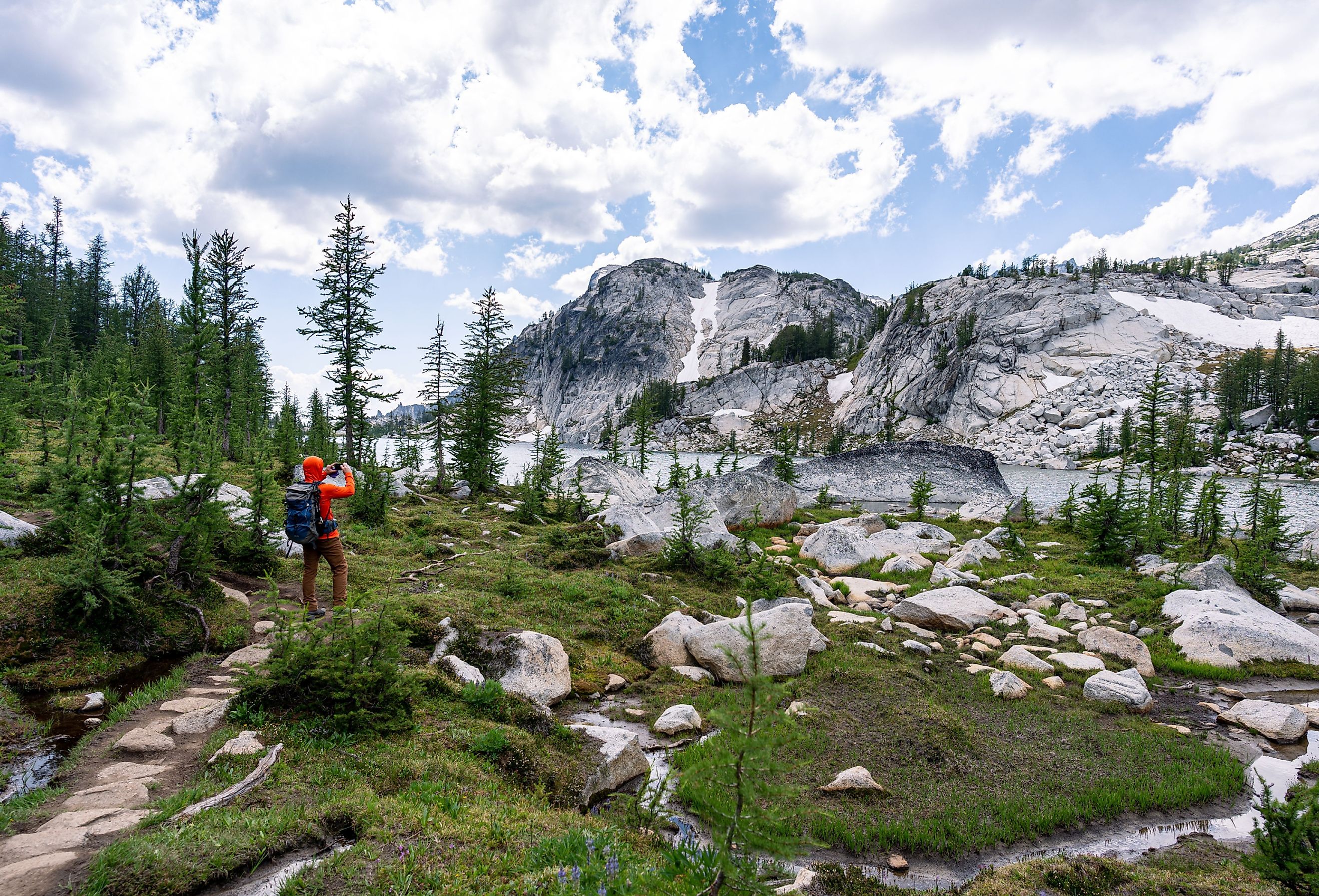 A man in bright orange shirt standing on hiking trail to take picture of beautiful lake and mountain while backpacking in the Enchantments, Leavenworth, Washington. Image credit Suchavadee via Shutterstock.